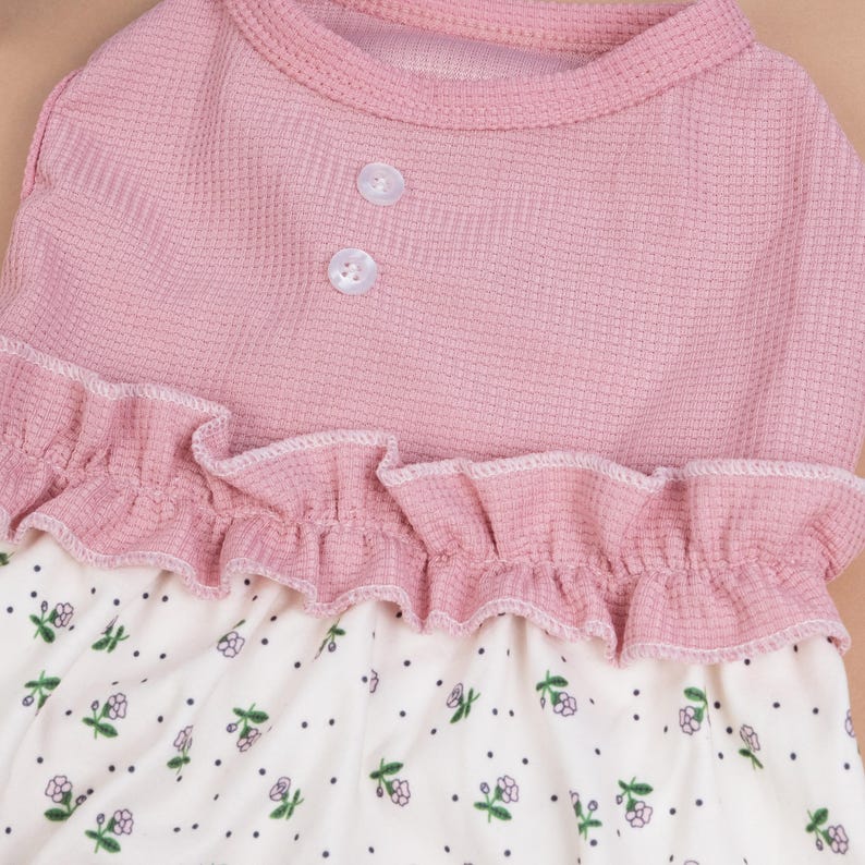 Peut inclure: Un v&ecirc;tement sans manches avec un corsage en maille gaufr&eacute;e rose poudr&eacute; dot&eacute; de deux petits boutons blancs &agrave; l'encolure. Un volant horizontal s&eacute;pare le corsage d'une jupe cr&egrave;me orn&eacute;e d'un motif de minuscules points noirs et de petites fleurs.