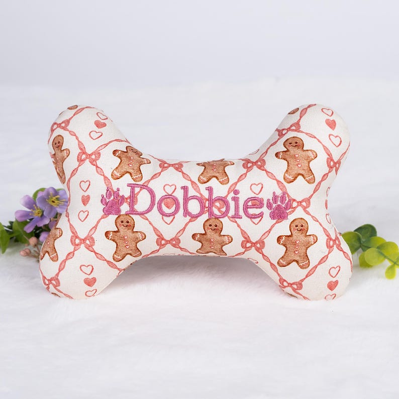 Op de afbeelding: Een botvormig hondenspeeltje met een witte achtergrond en een patroon van peperkoekmannetjes, hartjes en roze strikken. De naam "Dobbie" is roze geborduurd. Er is ook een pootafdruk op het speeltje geborduurd.