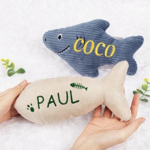 May include: Two plush toys shaped like fish. One is a blue shark with the name "Coco" embroidered on it. The other is a beige fish with the name "Paul" and a small green fishbone embroidered on it.