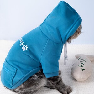 May include: A blue hooded sweatshirt for a cat, featuring a front pocket and a drawstring hood. The sweatshirt has the word "Smey" and a paw print embroidered on the back. A gray and white cat is wearing the sweatshirt. A gray toy with the name "PAUL" is next to the cat.