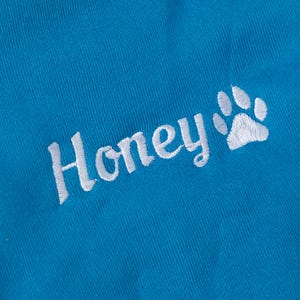 May include: Close-up of a bright blue fabric with the embroidered word "Honey" in white script, accompanied by a white paw print. The fabric has a textured appearance.