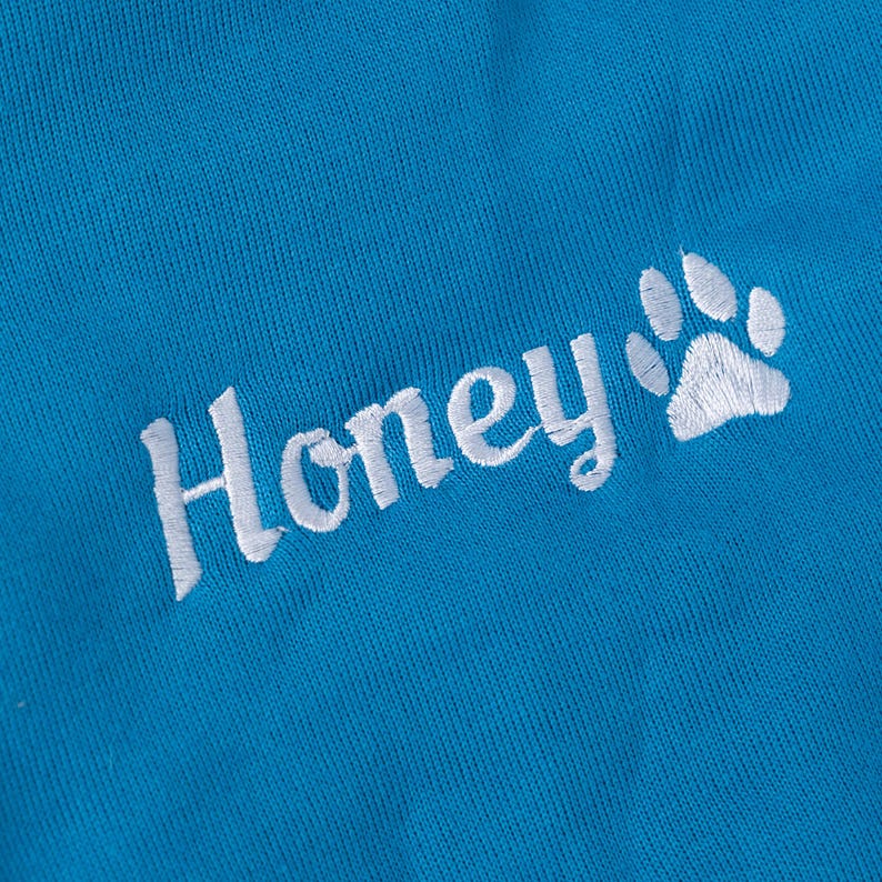 May include: Close-up of a bright blue fabric with the embroidered word "Honey" in white script, accompanied by a white paw print. The fabric has a textured appearance.