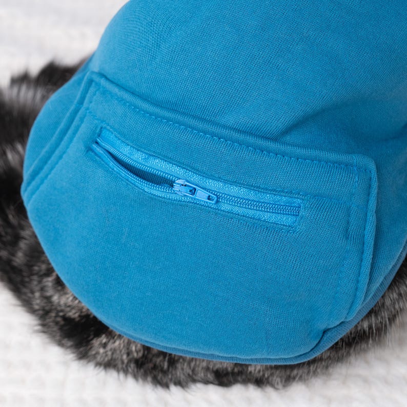 May include: Close-up of a turquoise blue dog jacket with a zippered pocket. The jacket is made of a soft, textured fabric. The zipper is also turquoise blue. The jacket is resting on a bed of gray and white faux fur.