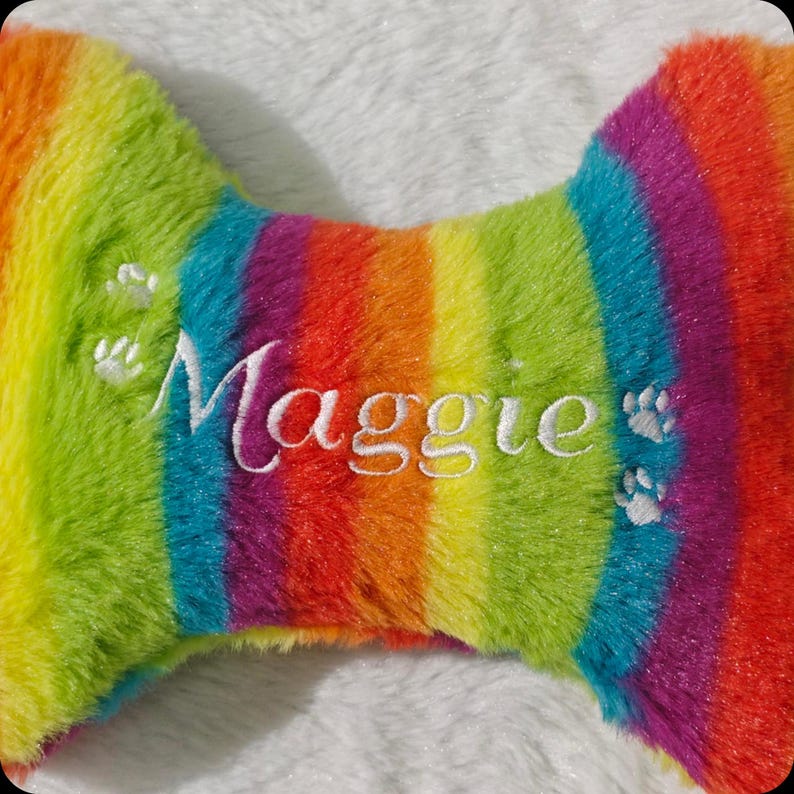 Op de afbeelding: Een regenboogkleurig pluche hondenbot speelgoed met de naam "Maggie" in het wit geborduurd. Het speelgoed heeft pootafdrukken en is gemaakt van zacht, pluizig materiaal.