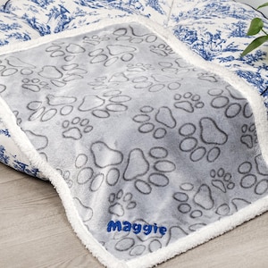 May include: Gray dog bed blanket with a white sherpa border. The blanket has a paw print pattern and the name "Maggie" embroidered in blue on the bottom.