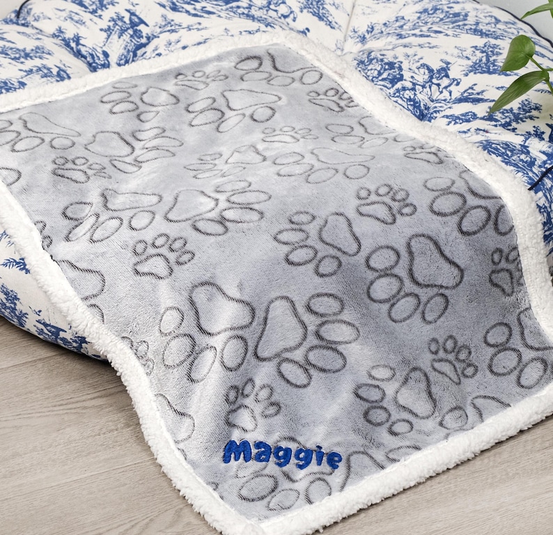 May include: Gray dog bed blanket with a white sherpa border. The blanket has a paw print pattern and the name "Maggie" embroidered in blue on the bottom.
