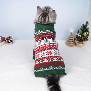 May include: A cat wearing a green, red, and white Christmas sweater. The sweater has a pattern of snowflakes, trees, and zigzags. The name "Luna" is embroidered on the back. The cat is facing away from the camera.