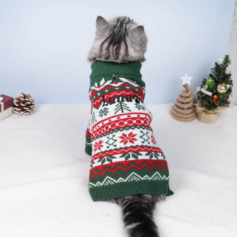 May include: A cat wearing a green, red, and white Christmas sweater. The sweater has a pattern of snowflakes, trees, and zigzags. The name "Luna" is embroidered on the back. The cat is facing away from the camera.