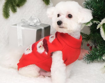 May include: A white dog wearing a red Christmas sweater with a snowman design. The dog is sitting on a white fluffy surface with a silver gift box in the background.
