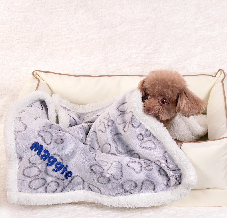 May include: A gray and white dog bed blanket with a paw print design. The blanket has a white border and the name "Maggie" embroidered in blue on the blanket. A small brown dog is curled up on the blanket.