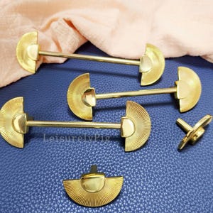 May include: Gold-coloured metal hardware pieces with a fan-shaped design. The hardware includes several long pieces and a few smaller components, all with a shiny finish. The items are arranged on a blue surface with a peach-coloured fabric in the background.