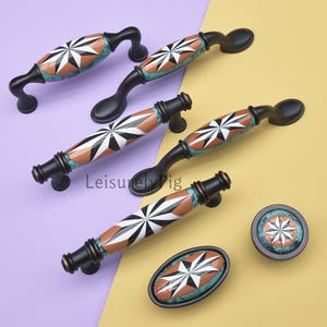 Può includere: Una collezione di maniglie e pomelli neri per armadi con un design a stella. Le maniglie hanno una base nera con un centro in ceramica con un motivo a stella bianco, nero e arancione. I pomelli hanno un design simile. Lo sfondo è viola e giallo.