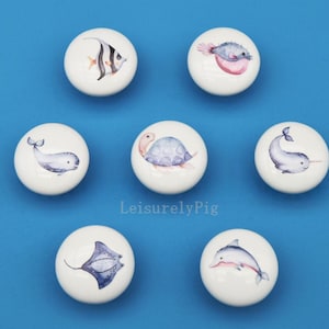 Puede incluir: Siete pomos de cerámica blanca con ilustraciones de acuarela de criaturas marinas, incluyendo un pez, una tortuga, una ballena, un narval, una manta raya y un delfín. Los pomos están sobre un fondo azul. El texto "LeisurelyPig" es visible.