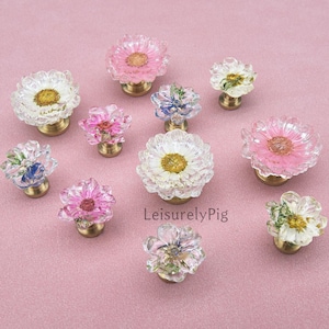 Puede incluir: Un conjunto de tiradores decorativos en forma de flor, en varios colores, incluyendo rosa, blanco y azul. Cada tirador presenta un diseño floral transparente y texturizado con una base dorada. Los tiradores están dispuestos sobre una superficie rosa.