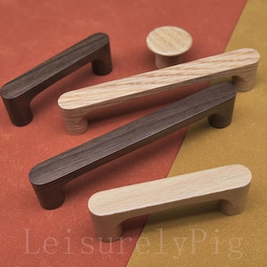 Może przedstawiać: Asortyment drewnianych uchwytów do szafek i gałka w różnych odcieniach brązu i naturalnego drewna. Uchwyty są prostokątne z zakrzywionymi uchwytami. Widoczny jest tekst "LeisurelyPig".