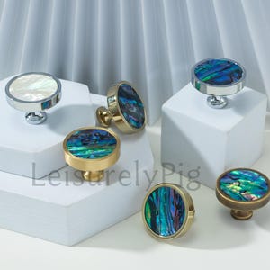 Op de afbeelding: Een verzameling decoratieve kastknoppen met iriserende abalone schelpinleg. De knoppen hebben verschillende metalen afwerkingen, waaronder zilver en goud, en worden gepresenteerd op witte geometrische platforms. De knoppen zijn rond en gepolijst.
