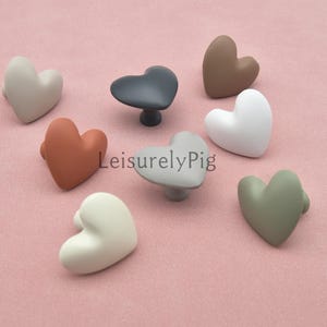 May include: A collection of heart-shaped cabinet knobs in various colors, including white, gray, brown, and green. The knobs are arranged on a pink surface, with the text "LeisurelyPig" visible.