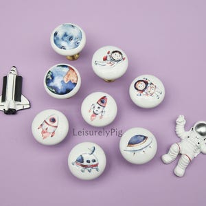 Puede incluir: Una colección de pomos de armario y elementos decorativos con temática espacial. Los pomos presentan diseños de planetas, cohetes y astronautas. También hay un transbordador espacial blanco y negro y una figura de astronauta blanca. El fondo es de color lavanda.