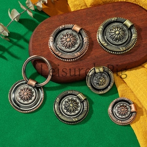 Peut inclure: Ensemble de boutons et poignées de meubles de style antique en bronze et cuivre. La quincaillerie présente un motif floral au centre, entouré d'une bordure décorative. Certaines poignées ont une poignée en anneau. Les articles sont présentés sur une surface en bois.