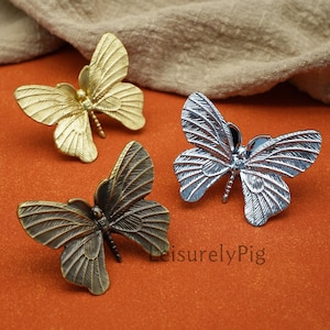 Op de afbeelding: Drie metalen decoratieve vlinderaccenten in goud, zilver en brons. Elke vlinder heeft gedetailleerde vleugelpatronen en een getextureerd lichaam. De vlinders zijn gerangschikt op een oranje oppervlak met een neutraal gekleurde stof op de achtergrond.