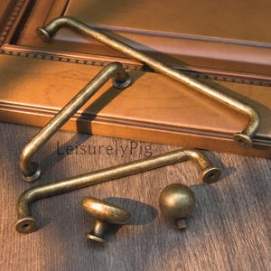 May include: A collection of antique brass cabinet hardware, including bar pulls and round knobs. The hardware is displayed on a wooden surface, showcasing the aged finish and design. The text "LeisurelyPig" is visible.