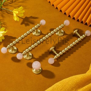 May include: A collection of brass cabinet handles and a knob, each featuring a pink quartz sphere. The handles have a beaded design and are mounted on circular bases. The hardware is displayed on a mustard-coloured surface.