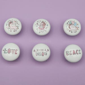Peut inclure: Six boutons de placard en céramique blanche avec des motifs colorés de licornes et de texte. Les boutons présentent des illustrations de licornes, une ballerine et les mots "Love", "Be Like A Unicorn" et "Peace". Les boutons sont posés sur un fond violet.