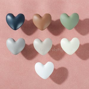 以下が含まれることがあります： ネイビー、ブラウン、セージグリーン、グレー、オフホワイト、ホワイトなど、さまざまな色のハート型のキャビネットノブ7個セット。 ピンク色の表面に配置され、それぞれが影を落としています。 メートル法が適用されます。