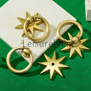 Puede incluir: Cuatro tiradores de latón para armarios con diseño de estrella. Cada tirador presenta un asa circular unida a una base en forma de estrella. Los tiradores son de un tono dorado cálido, con la palabra "Leisurely" visible en la imagen.