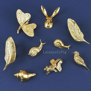 Może przedstawiać: Kolekcja małych, złotych przedmiotów dekoracyjnych, w tym motyl, głowa królika, pióra, ślimaki, muszla, ryba i ptak. Przedmioty są ułożone na niebieskiej powierzchni. Widoczny jest tekst "LeisurelyPig".