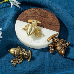 Pode incluir: Três puxadores de armário antigos dourados em forma de bolota com detalhes de folhas. Um puxador está preso a uma superfície redonda de madeira e mármore branco. Os puxadores são um elemento decorativo de hardware.