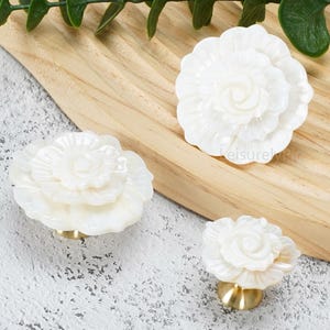 May include: Three white flower-shaped cabinet knobs with layered petals and a glossy finish. Each knob features a gold-toned metal base. The floral designs vary in size, showcasing intricate, carved-style petal details on each individual piece.