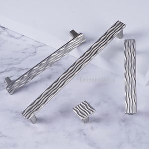 May include: A collection of silver-toned cabinet hardware, including two long pulls, a small square knob, and a rectangular pull. The hardware features a textured, wavy design. The items are arranged on a white and gray marble surface.
