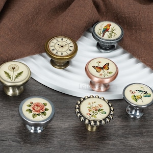 Op de afbeelding: Een verzameling van zeven ronde kastknoppen met metalen voetjes in diverse afwerkingen. Elke knop heeft een unieke ronde inzet met botanische, dierlijke of klokillustraties, waaronder een roos, een vlinder, een papegaai en een paardenbloem.
