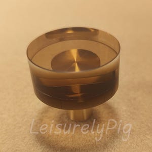 Puede incluir: Un tirador decorativo para armario con una parte superior transparente de color ámbar y una base de color latón. El tirador tiene un diseño circular y está sobre un fondo neutro. El texto "LeisurelyPig" es visible.