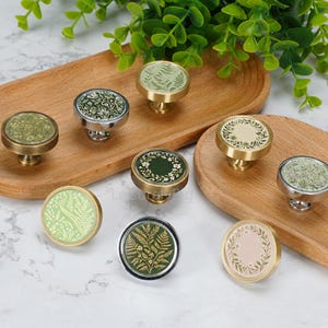 May include: A collection of eight round cabinet knobs featuring various botanical designs, including fern leaves and floral wreaths, set in metallic gold and silver finishes. The knobs are arranged on two wooden trays with a marble-patterned surface.