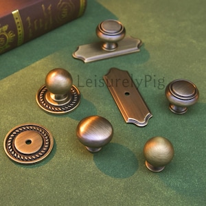 Può includere: Una collezione di ferramenta per mobili in ottone antico, tra cui pomelli e maniglie. L'hardware presenta vari design, come pomelli rotondi, pomelli con bordi a corda e maniglie rettangolari. L'hardware ha una finitura calda e invecchiata.