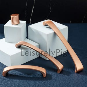 May include: Copper-colored cabinet hardware set, including handles and a cylindrical knob. The handles have a curved design and are displayed on white blocks against a dark blue background. The text "LeisurelyPig" is visible.