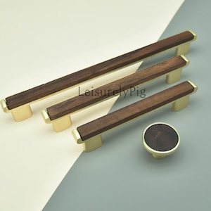 May include: A set of four cabinet hardware pieces: three rectangular pulls and one round knob. The pulls have a dark brown wood center and gold-colored metal ends. The knob has a matching wood center and gold base. The text "LeisurelyPig" is visible.