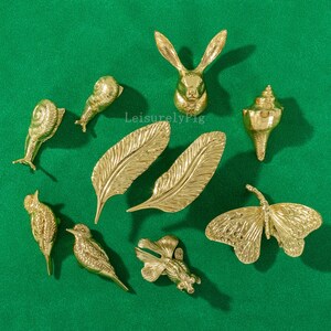 Może przedstawiać: Kolekcja złotych przedmiotów dekoracyjnych, w tym królik, ślimak, pióra, muszla, ptak, ryba i motyl, ułożonych na zielonej powierzchni. Obiekty mają skomplikowane detale i metaliczne wykończenie. Widoczny jest tekst "LeisurelyFig".