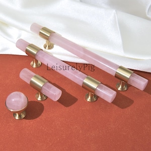 Puede incluir: Un conjunto de herrajes para armarios de cuarzo rosa, que incluye dos tiradores largos, un tirador más corto y un pomo redondo. Los herrajes presentan un centro de piedra rosa pálido con detalles y bases de metal dorado. Los artículos están dispuestos sobre una superficie marrón rojizo.