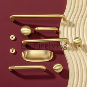 Pode incluir: Uma coleção de ferragens douradas para armários, incluindo puxadores, botões e puxadores, dispostos sobre uma superfície bordô. As ferragens variam em tamanho e formato, com uma mistura de designs arredondados e em barra. A palavra "LeisurelyPig" é visível.