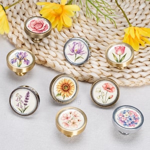 Puede incluir: Un conjunto de ocho pomos redondos para armarios con diversas ilustraciones florales, incluyendo una rosa, un iris, un tulipán, un girasol, una amapola, lavanda, flores de cerezo y una hortensia, en marcos metálicos de tonos dorados y plateados.
