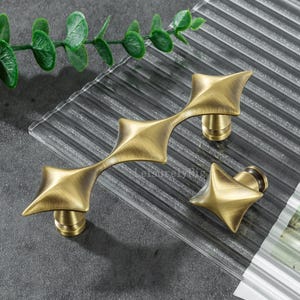 May include: Antique brass cabinet hardware set. The set includes a long handle and a single knob, both with a starburst design. The hardware is displayed on a textured glass surface, with a sprig of green leaves in the background.