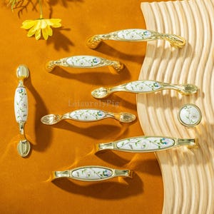 Könnte beinhalten: Eine Sammlung goldfarbener Schrankgriffe und -knöpfe mit weißen Keramikmitteln. Die Keramik weist ein zartes Blumenmuster in Blau, Grün und Lila auf. Die Hardware ist auf einer Holzoberfläche und einem senffarbenen Hintergrund angeordnet.