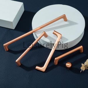 Puede incluir: Juego de herrajes para armarios de color cobre, que incluye manijas de varios tamaños y un pomo. Las manijas tienen un diseño moderno y minimalista con forma curva. El hardware se exhibe sobre una plataforma circular blanca y una superficie azul oscuro.