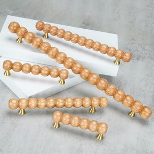 May include: A collection of five cabinet pulls featuring a light brown wood grain finish with a spherical, beaded design. Each pull is supported by two small, conical brass-toned metal mounts that attach to the ends of the wooden handle.