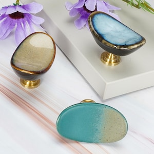 May include: Three cabinet knobs with irregular, pebble-like shapes and glossy, multi-coloured glazed finishes. One knob is beige and brown, one is blue and black, and one is teal and beige. Each knob features a polished gold-tone metal base.