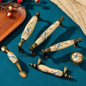 Pode incluir: Um conjunto de puxadores de armário de cerâmica e um botão. Os puxadores têm um fundo creme com um desenho de pássaro azul e floral. A ferragem é de cor bronze. O botão tem um design correspondente. Os itens estão dispostos em uma superfície azul-petróleo.