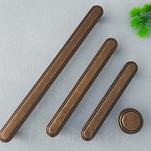 Peut inclure: Un ensemble de quincaillerie de meubles de couleur bronze, comprenant trois poignées allongées et un bouton rond. La quincaillerie présente un design rectangulaire avec des extrémités arrondies et un bord rainuré. Les articles sont disposés en diagonale sur une surface gris clair.